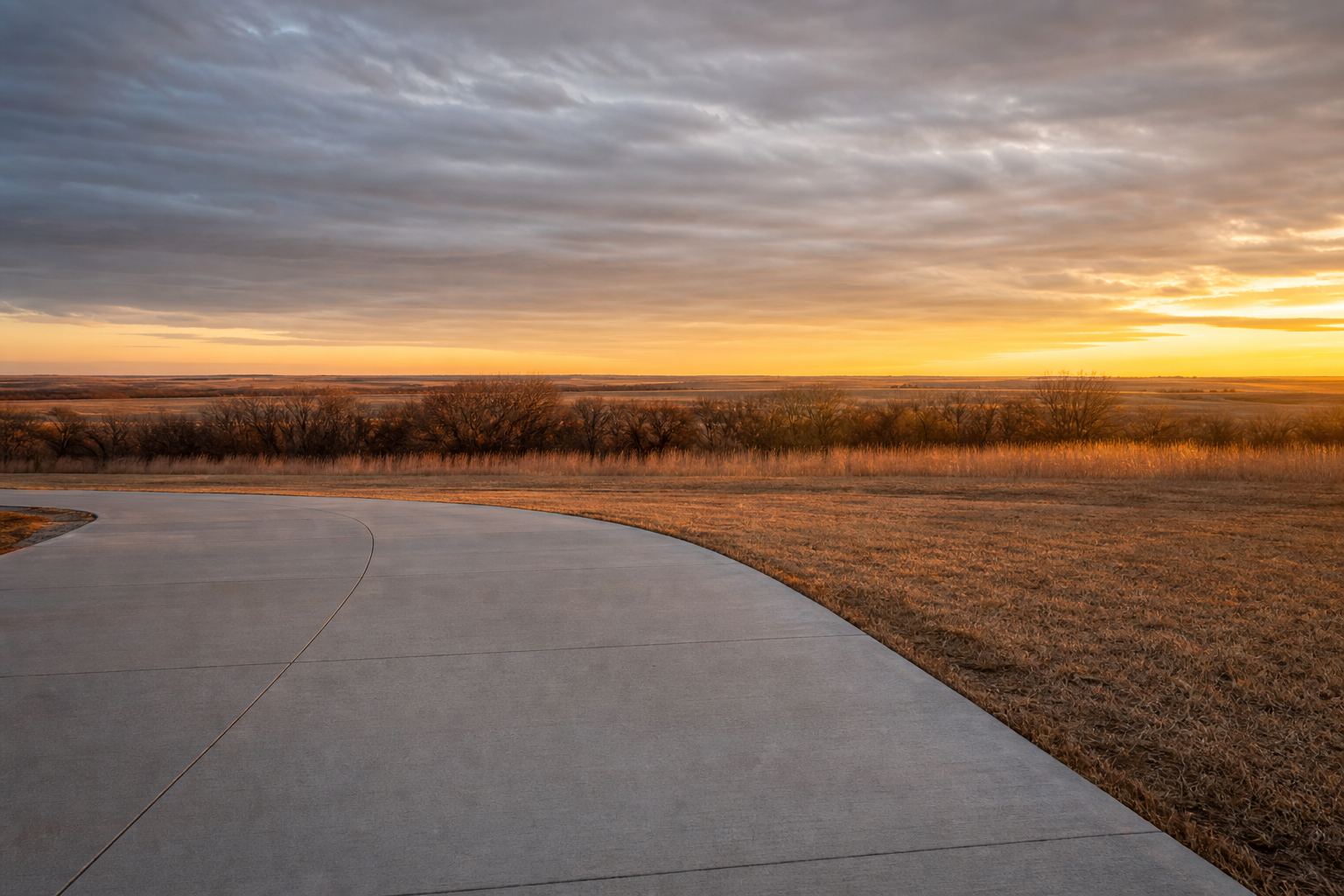 Curved concrete driveway entrance beside open prairie at sunset